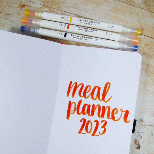 Narrow travelers journal open to a page with ‘meal planner 2023’ written on it and calliograph pens 