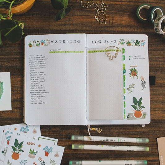 A flatlay photo of a journal open on a dark background surrounded by green and plant-related journaling supplies. The spread is showing a plant care watering log for watering your houseplants.