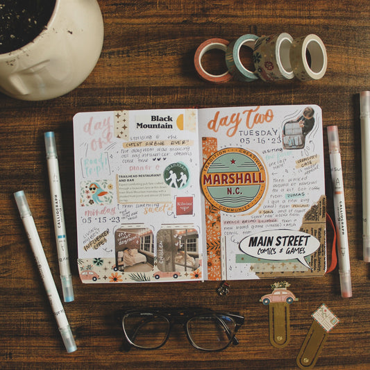 A dot grid notebook is lying open on a dark table. On the pages is a travel memories spread using washi tape, photos, lettering, and cut out trip memorabilia. The notebook is surrounded by various stationery items.