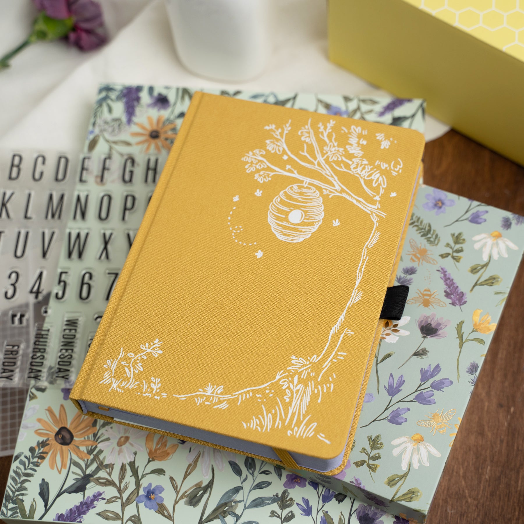 Yellow notebook with white Bee and floral design on a wooden surface with other notebooks and stationery items.